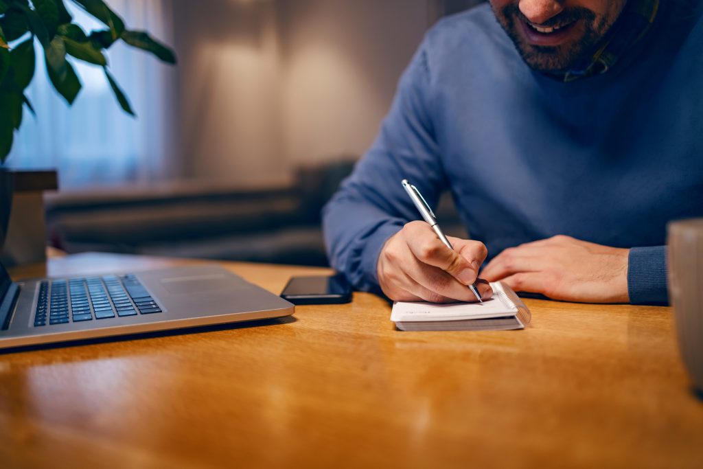 Define Goals, Write Tasks, Do A Checklist. A Happy Young Businessman Is Sitting At Home, Working Remotely, And Writing Tasks In His Spiral Block. There Is A Laptop On A Table.